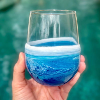 A hand holds the Artful Nests Resin Wave Stemless Wine Glass, showcasing blue and white swirls that evoke ocean waves—a perfect piece of beach glassware.