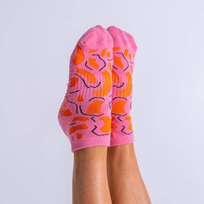 A pair of feet raised, wearing Reformer Loops Pop Chaos Pilates Ankle Grip Socks in pink with bright orange and black abstract patterns, set against a plain light background.