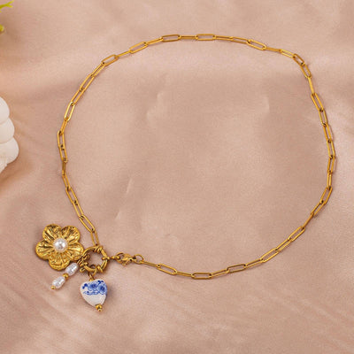 The Delphine Charm Necklace by JOSSLYN by wall to wall features a tarnish-free gold chain with a large flower charm, a blue and white porcelain heart charm, and pearl accents, shown on beige fabric.