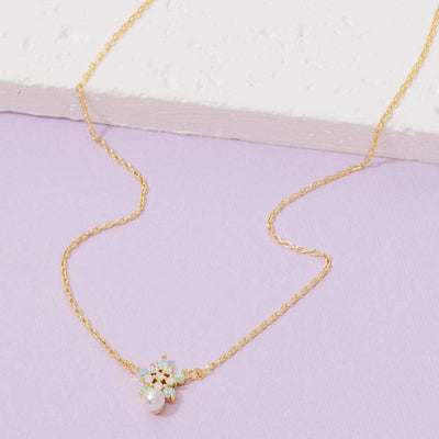 The Hermia Pendant Necklace by Fame Accessories features a delicate gold-dipped chain with a cubic zirconia flower pendant adorned with light blue and white stones and a dangling pearl, shown on a light purple and white background.