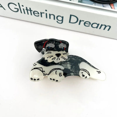 The Schnauzer Hair Claw Clip by SoapElenGlen, featuring a black and white dog design, rests on a white surface beside fashion jewelry and part of a closed book titled A Glittering Dream.