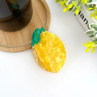 A lemon-shaped Acetate Yellow Lemon Hair Claw Clip by The Diva Soap sits on a wooden surface. Bright yellow flowers and a leafy plant frame the upper right corner.