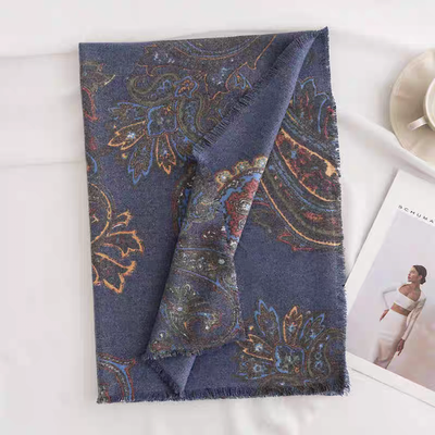 The Eugenia Paisley Print Scarf by Pretty Persuasions, folded to show its vibrant blue paisley pattern, rests on a white surface beside a cup, saucer, and a magazine displaying a woman in a white outfit.