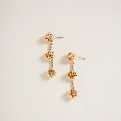 The Christmas Bows & Bling Earrings in Gold by Golden Stella are 2-inch gold dangle earrings with three floral clusters linked by sparkling crystals, crafted from zinc in China, displayed on a plain off-white background.
