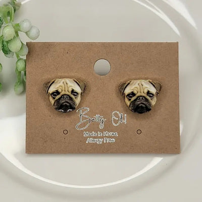 A pair of Pug Face Enamel Post Earrings from BETTYOH INC is displayed on a brown card labeled BETTYOH INC Made in Korea Allergy Free, resting on a white plate with a green plant nearby.
