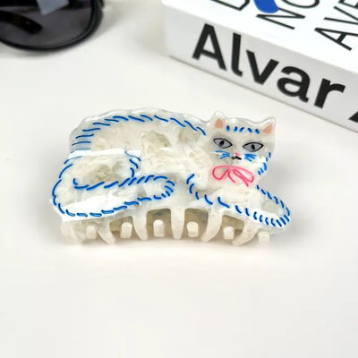 The Acetate Cute Persian Cat Hair Clip by The Diva Soap, crafted from lightweight material and shaped like a white cat with blue stitching and a pink bow, rests on the table next to sunglasses and a partially visible book titled Alvar.