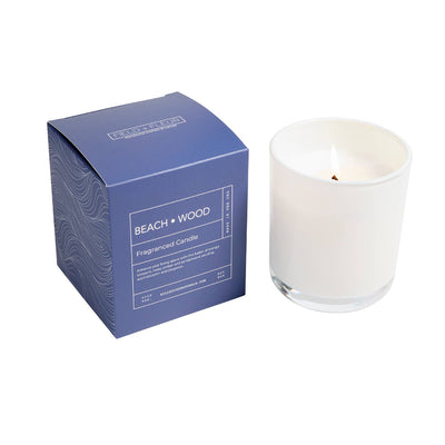 A white candle in a clear glass holder sits beside a blue Beach Wood Boxed Glass Candle (9oz.) by Hillhouse Naturals/Field+Fleur, with subtle sandalwood notes, on a white background.