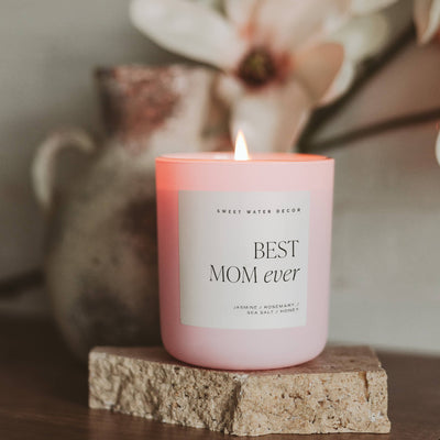 A pink Best Mom Ever soy candle by Sweet Water Decor, infused with jasmine, rosemary, sea salt, and honey oils, burns gently on a stone coaster. It sits against a blurred backdrop with a ceramic vase and softly focused flowers.