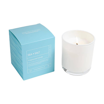 A white Hillhouse Naturals/Field+Fleur Sea Salt Boxed Glass Candle (9oz) glows beside a light blue box. With driftwood and ocean air notes, its minimalist design creates a refreshing, coastal atmosphere.