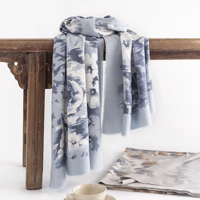 A Pretty Persuasions Floral Escape Print Scarf in light blue with a white and gray floral pattern is draped over a wooden table, accompanied by matching folded fabric and a beige cup and saucer nearby.