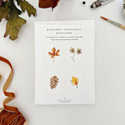 The Fall Leaves Paintable Notecards by emily lex studio features four autumn watercolor designs—maple, multi-lobed, oak, and yellow leaf—perfect for DIY greeting cards. Art supplies, ribbon, and dried leaves add creative flair.