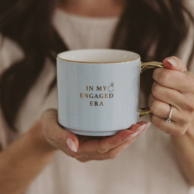 In the persons hand is the In My Engaged Era fine bone china mug by Sweet Water Decor, featuring elegant blue detailing, a golden handle, and a small ring graphic that beautifully complements the engagement ring on their finger.