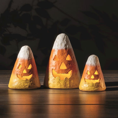 Mud Pie’s Candy Corn Luminaries—three candy corn-shaped papier-mâché decorations with jack-o-lantern faces—glow with yellow flameless tea lights (batteries included) on a wooden surface, set against a dark background.