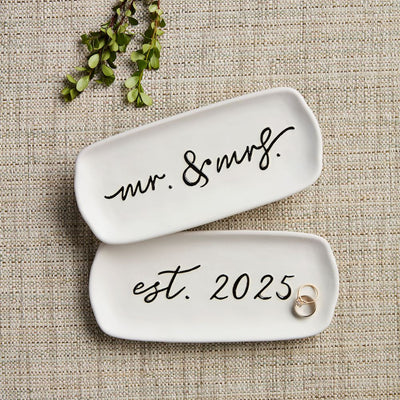 Two white hand-painted stoneware trays from Mud Pies Wedding Everything Dishes rest on a textured surface. One reads mr. & mrs. and the other est. 2025, holding a pair of rings, with a sprig of greenery above—ideal for special moments.