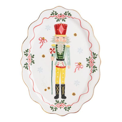 The Mud Pie nutcracker platter is a ceramic, scalloped-edge tray featuring a painted nutcracker with a candy cane and festive red, green, and gold holiday motifs like bows, holly, and snowflakes.