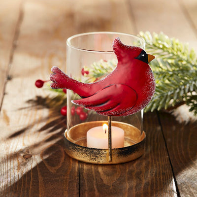 The Mud Pie Cardinal Votive features a red tin cardinal on a clear glass holder with a gold metal base, displaying a lit tealight. Pine branches and red berries accent the wooden surface nearby.