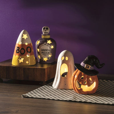 Mud Pie Halloween Votive Sets decorate a table with glowing ceramic pieces: a hand-painted candy corn with BOO, a purple skull poison bottle, a ghost, and a jack-o-lantern witch, each lit by flameless tea lights.