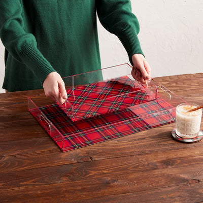 A person in a green sweater picks up a Mud Pie Tartan Acrylic Tray (UNAVAILABLE FOR SHIPPING) from a matching placemat on a wooden table, while a glass of creamy drink with cinnamon sits on a plaid coaster nearby.