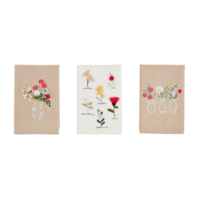 Mud Pies Flower Embroidery Towels set features three tea towels: the left and right, embroidered with floral designs and French knot details on chambray, while the center white towel displays botanical illustrations with labeled text.