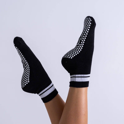 A person's legs raised with feet up, wearing Reformer Loops Quarter Pilates Grip Socks in black with white silicone soles and striped cuffs, shown against a plain light backdrop.
