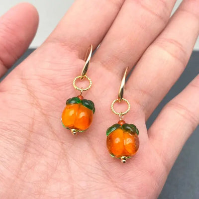 A hand holds the Glazed Persimmon Dangle Earrings by Mio Queena, featuring gold-plated accents with orange fruit-shaped charms and green leaves, resembling tiny pumpkins.