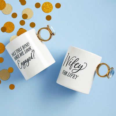 The Wedding Ring Mugs by Mud Pie include two white ceramic mugs with hand-painted gold and blue gemstone ring-shaped handles. One mug reads Does this ring make me look engaged? and the other says Wifey for Lifey, all set on a light blue background with gold confetti.