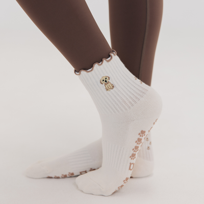 A person wearing Almla Active Golden Pilates Socks—white ribbed socks with an embroidered Golden Retriever near the ankle and brown paw print grips—stands on a light background. The perfect dog lover gift.