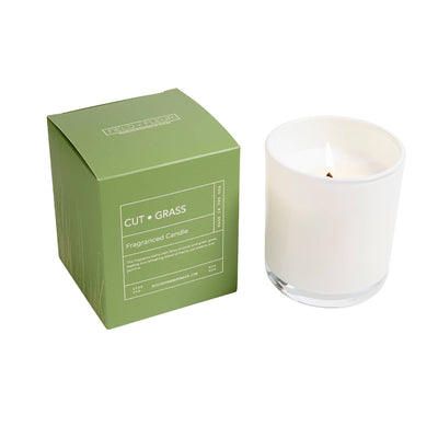 A lit Hillhouse Naturals/Field+Fleur Cut Grass Boxed Glass Candle (9oz) in a clear holder stands by its green, minimalist box. The candle releases a fresh scent with jasmine hints, and the box is labeled with the product name.