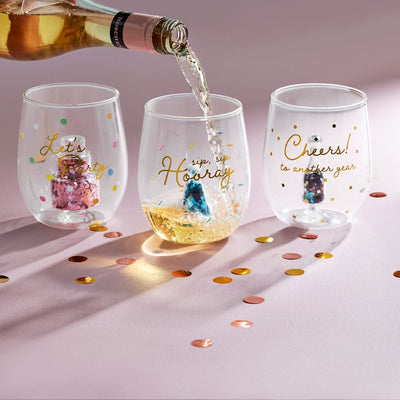 On a pink surface, three Mud Pie stemless wine glasses from the Birthday Icon Wine Glasses set display festive designs. One is being filled with sparkling rosé and reads sip, sip, Hooray. The others say Lets Party and Cheers! to another year, surrounded by confetti and glitter.