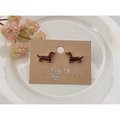 Chocolate Dachshund Post Earrings by BETTYOH INC, shown on a branded brown card atop a white plate with pink dried flowers. These cute earrings feature titanium posts and are lead & nickel free.