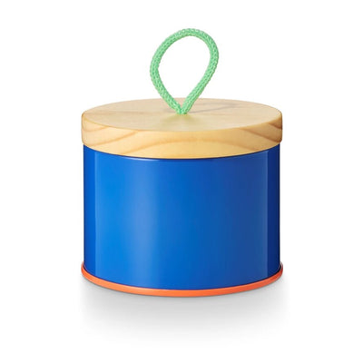 The Beach Towel Sun Tin Candle by Illume, a cylindrical blue container from Illume Candles, features a wooden lid with a green loop handle. It has an orange bottom rim, contrasting elegantly with the blue body and natural wood lid, set against a white background.