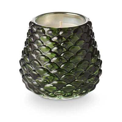 The Balsam & Cedar Pinecone Votive Candle by Illume Candles features a dark green, textured glass pinecone holder with a metallic interior and unlit wick, displayed against a white background.