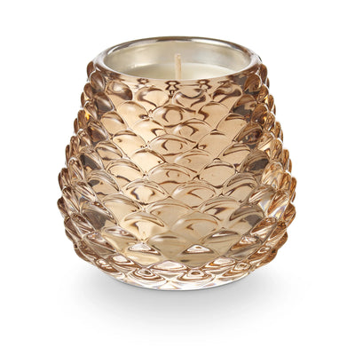 The Illume Candles Woodfire Pinecone Votive Candle features hints of smoky cedar in a textured, translucent glass holder with a gold-tinted, scale-like pinecone design, shown against a white background.