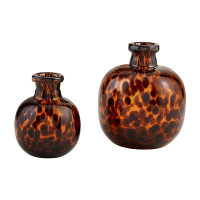 The Tortoise Glass Vases from Mud Pie feature a striking tortoiseshell pattern against a white background. The smaller vase is round with a narrow neck, while the larger one is elongated with broader dimensions in rich brown and amber hues.