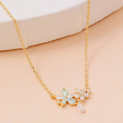 The Irina Necklace by Fame Accessories features a gold-dipped chain with a cubic zirconia flower pendant in light blue, clear, and white stones. Displayed on a peach surface beside a textured white object. MADE IN KOREA.