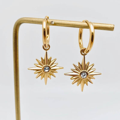 The Mio Queena Shooting Star Pendant Huggies feature gold hoops with dangling starburst charms, each set with a sparkling cubic zirconia, displayed on a minimalist gold stand against a plain white background.