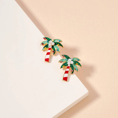 The Golden Stella Christmas Palm Tree Pearl Earrings feature red and white striped trunks, green glittery leaves, and pearl accents in a small post design, shown on a light beige background.