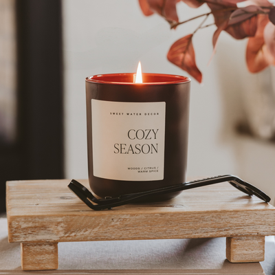 The Sweet Water Decor Cozy Season 15 oz Candle in a matte jar sits on a small wooden stand, with wick trimmers beside it. Warm light and autumn leaves in the background create a cozy, inviting atmosphere.