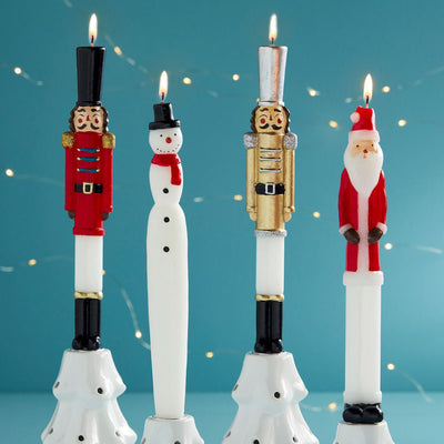 Four Mud Pie Christmas Figural Tapers—two nutcrackers, a snowman, and Santa Claus—are lit and displayed against a blue background with string lights.