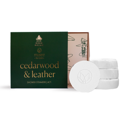 A green Musee Cedarwood & Leather Shower Steamers box with gold text sits open, revealing artwork inside. In front, three white, round shower steamers—infused with cedarwood essential oil—are stacked, one embossed with a leaf design.