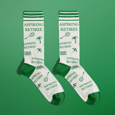 FUN CLUB’s Aspiring Retiree Socks are super-soft cotton with green toes, heels, and cuffs, featuring “Aspiring Retiree” text and fun tennis racket, palm tree, wave, and umbrella icons on a green background.