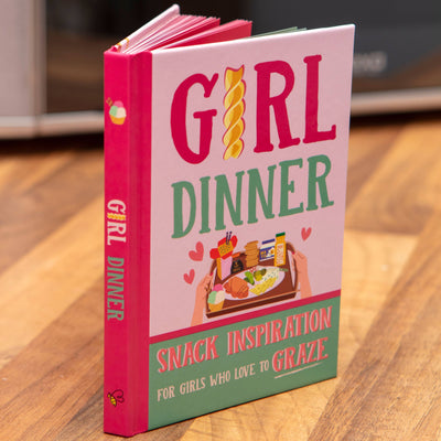 The Boxer Gifts Girl Dinner: 50 Small Snack Plate Ideas Recipe Gift Book stands upright on a wooden surface, its cover featuring playful snack illustrations and two hands holding a tray.