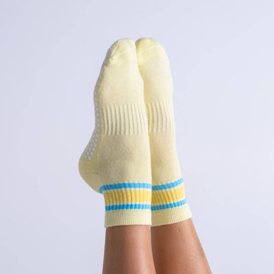 A pair of feet in Reformer Loops Cali Quarter Pilates Grip Socks—yellow with blue and yellow stripes at the ankles and white silicone soles—raised against a plain light background.