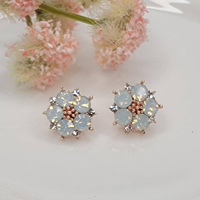 The Opal Flower Post Earrings by BETTYOH INC feature pale blue opal rhinestones and clear crystals in a gold-tone setting with titanium posts, shown on a white surface with pink dried flowers in the background.