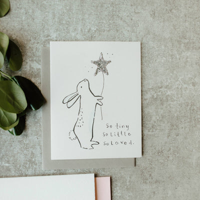 The So Tiny So Loved Baby Bunny Star Card by Sweet Julie Marie features a glittered line drawing of a bunny with a silver star balloon and the words “So tiny so little so loved”—a perfect congratulations card for new parents.