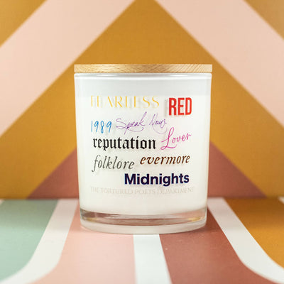 The Taylor Swift Albums Soy Candle by Unplug Soy Candles is a white soy candle in a glass jar with a wooden lid, showcasing album names in vibrant fonts—perfect for Swifties and displayed on a colorful, geometric background.