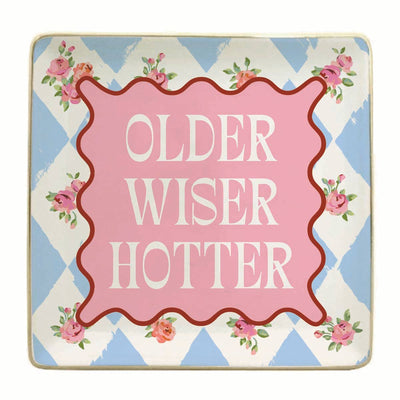 The soiree-sisters Older Wiser Hotter Ceramic Trinket Tray features a blue and white diamond pattern, pink corner flowers, a bold pink center with red border, and the phrase "OLDER WISER HOTTER"—a perfect decorative gift.
