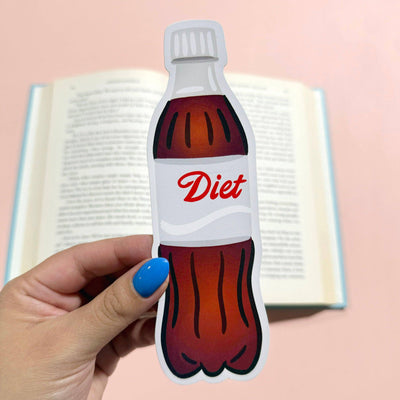 A hand with blue nail polish holds the Bloomwolf Studio Diet Soda Die Cut Bookmark, shaped like a soda bottle, in front of an open book on a pale pink surface.