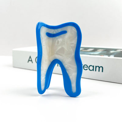 The SoapElenGlen White Tooth Acetate Hair Claw Clip, featuring a blue outline and white center, stands upright in front of a book on a white surface.