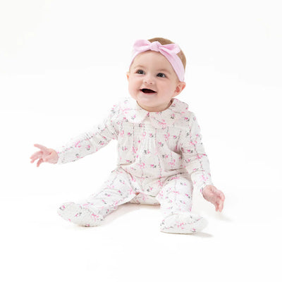 A smiling baby in the Dainty Buds and Bows Smocked Peter Pan Snap Footie by Angel Dear and a pink bow headband sits on a white background.